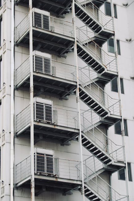 A tall, multi-storey building with external metal staircases on each level, featuring black treads and white railings, situated on a grey facade with several windows and air conditioning units. The st